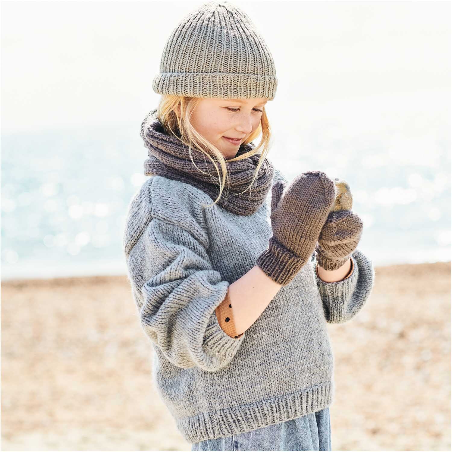 Essentials Super aran: Kind am Strand in grauem Strickpullover, Mütze und Schal von Rico Design.