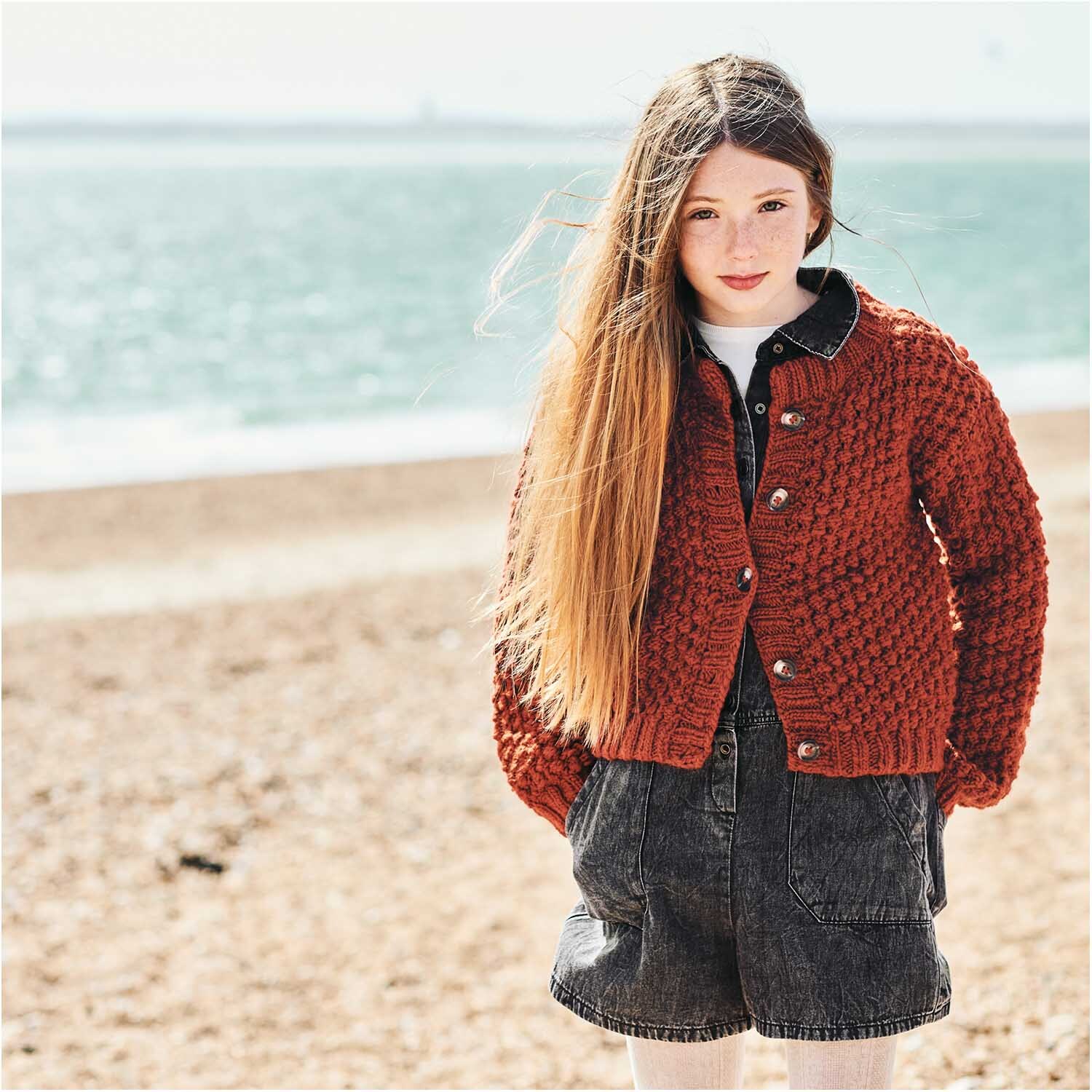 Essentials Super aran 50g 100m Farbe Rost von Rico Design Essentials Super aran: Mädchen am Strand trägt gestrickte Jacke in Rost von Rico Design, 50g 100m.