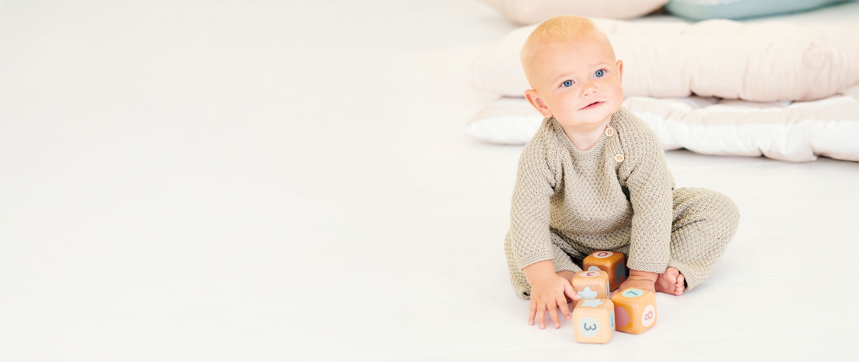 Baby sitzt mit Strickpulli und Strickhose auf Teppich.