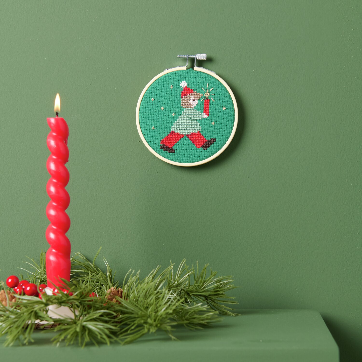 Stickpackung Lichterkind: Ein grüner Stickrahmen mit weihnachtlichem Motiv neben einer roten Kerze auf grünem Hintergrund.