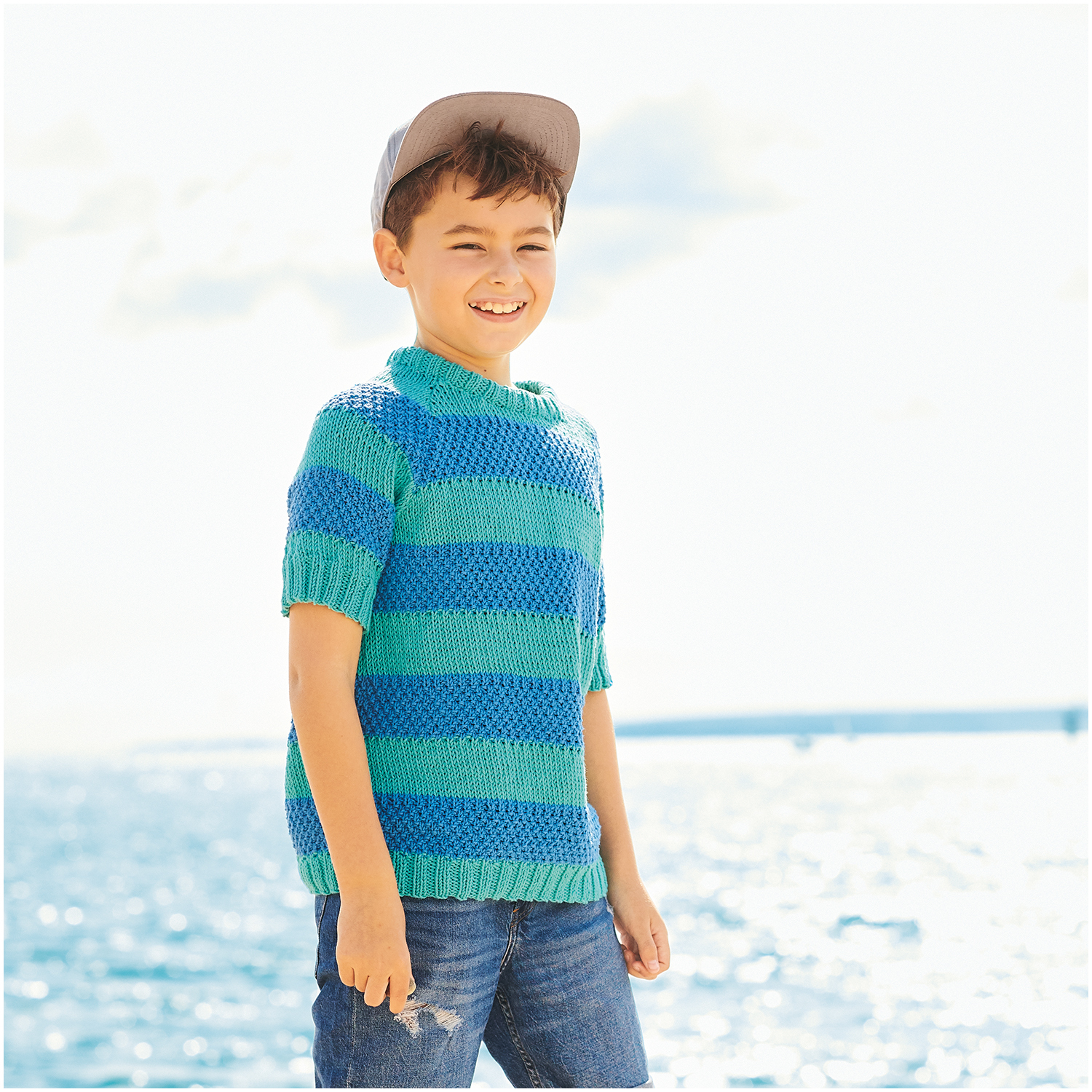 Essentials Organic Cotton aran: Junge in türkis-blauem Strickpullover am Meer, lächelnd, mit Cap.