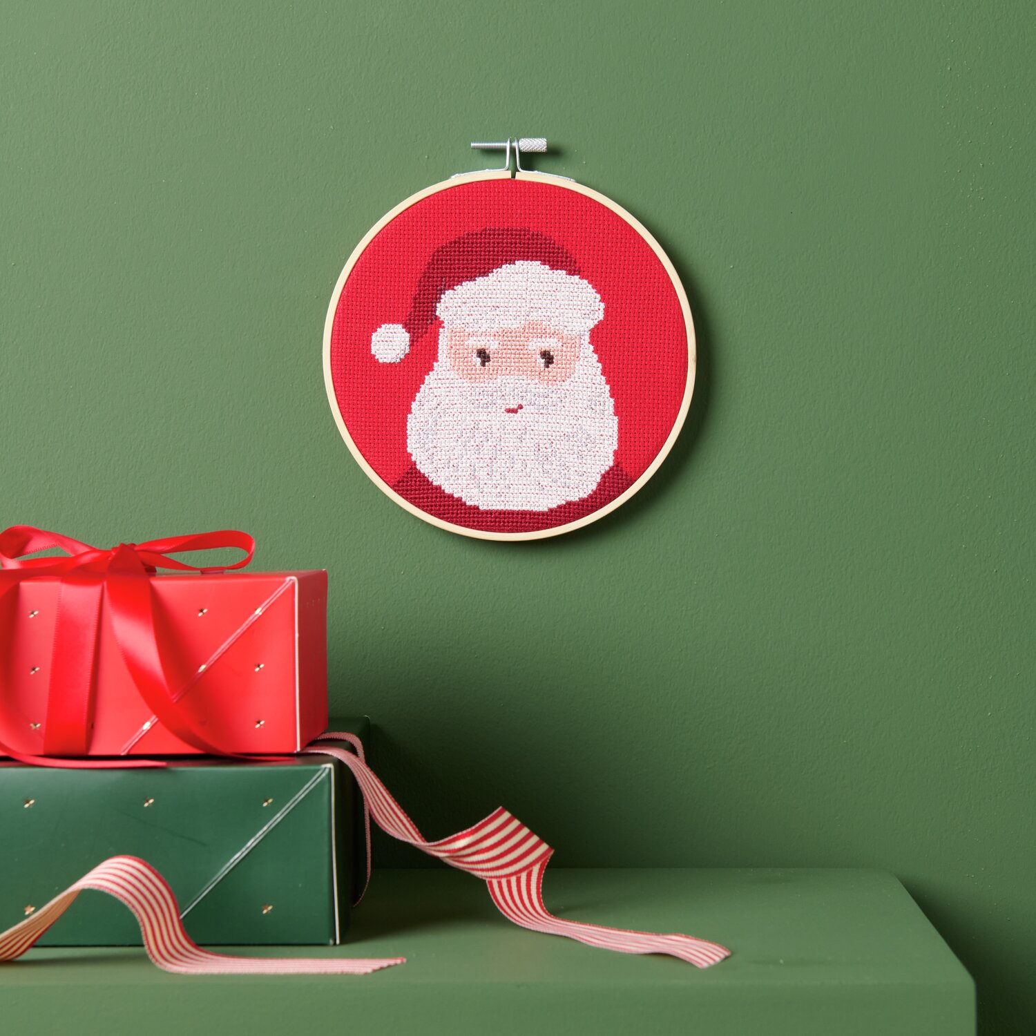 Stickpackung Portrait Santa zeigt ein gesticktes Weihnachtsmann-Gesicht auf rotem Hintergrund, aufgehängt an einer grünen Wand.