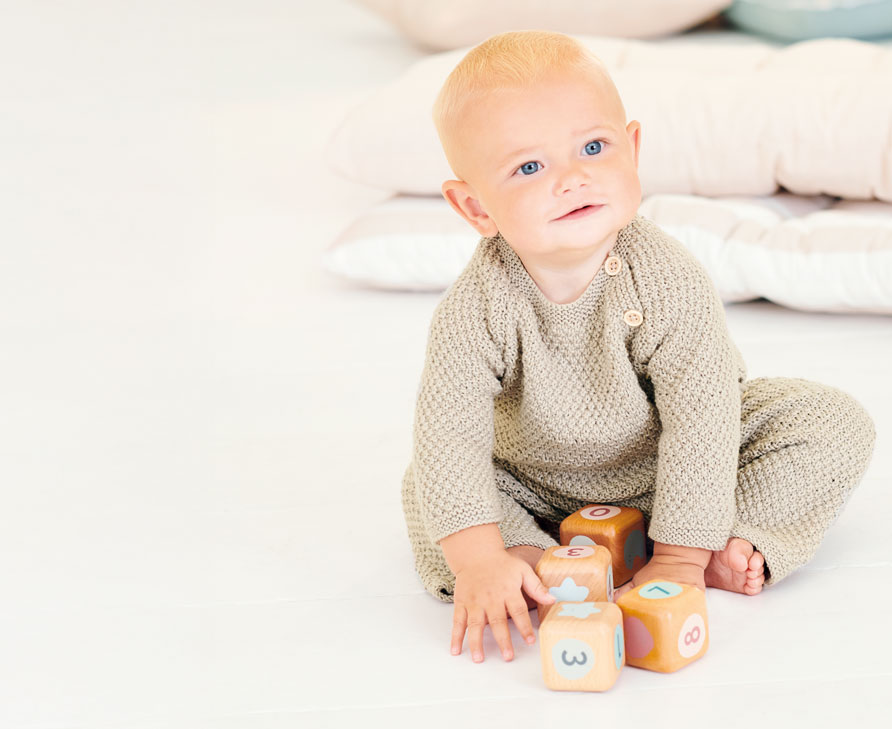 Baby sitzt mit Strickpulli und Strickhose auf Teppich.