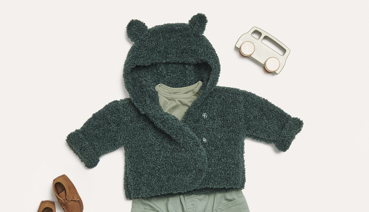Strickanleitung Babyjacke aus Creative Teddy: Flauschige, grüne Jacke mit Kapuze und Ohren, kombiniert mit Spielzeugauto und Babyschuhen.