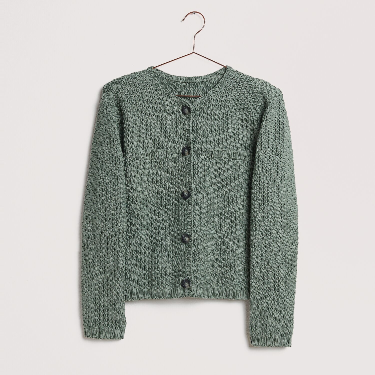 Strickset Jacke Modell 18 aus Die Neue Masche No. 12 von idee. Creativmarkt Strickset Jacke Modell 18 aus Die Neue Masche No. 12, in Salbei, auf einem Kleiderbügel präsentiert.