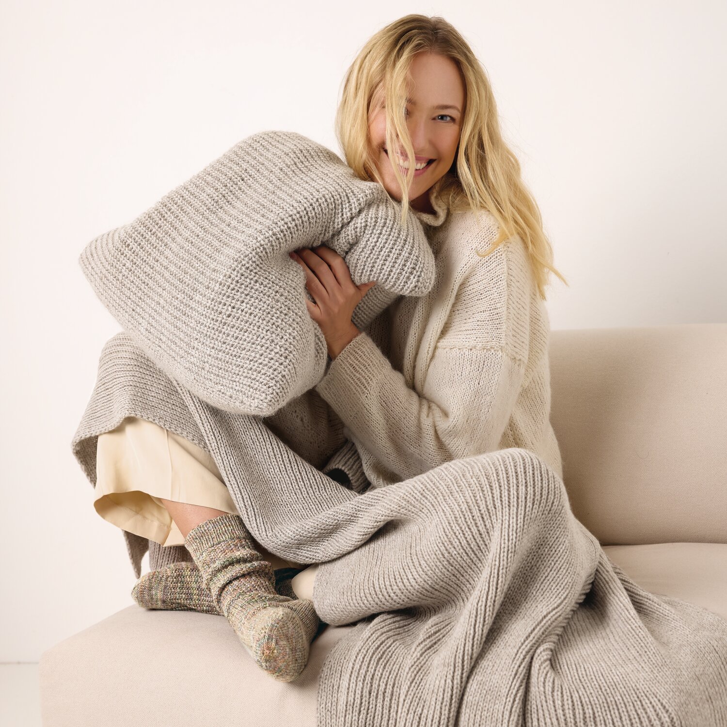 Strickset Kissen Modell 03 aus Die Neue Masche No. 13 von idee. Creativmarkt Strickset Kissen Modell 03: Frau in beigem Strickoutfit mit passendem Kissen auf Sofa.
