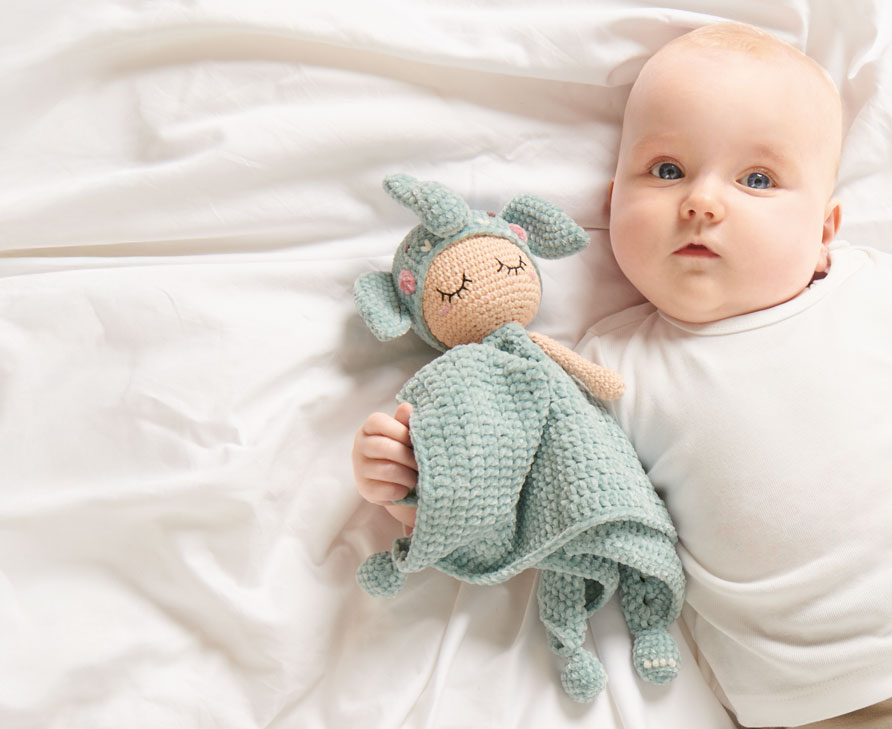 Ein liegendes Baby hält ein gehäkeltes Schnuffeltuch in Elefanten-Design im Arm. 
