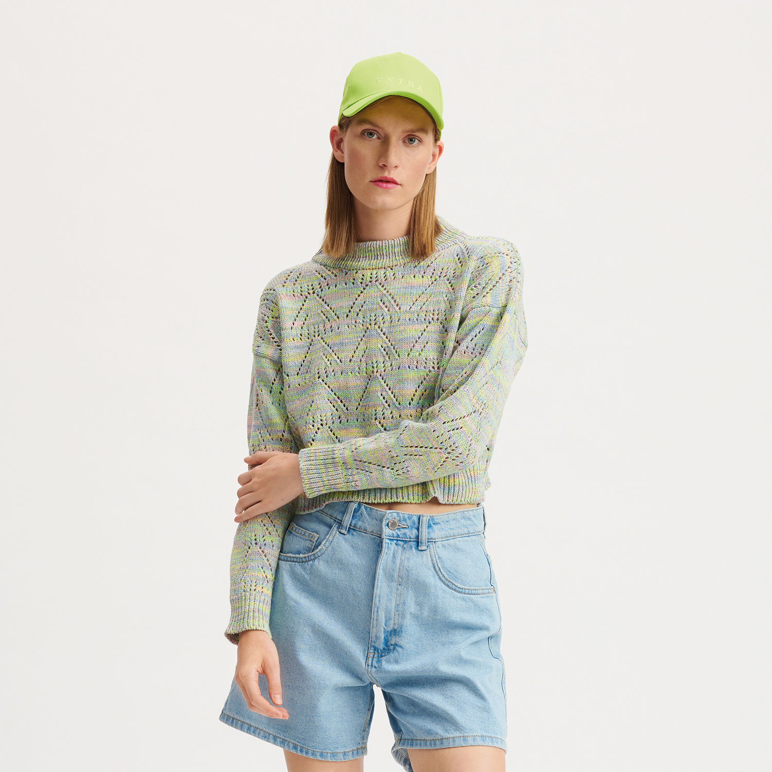 Strickanleitung Pullover aus Creative Summer Sprinkles: Frau trägt gestrickten Pullover in Pastellfarben, kombiniert mit blauer Jeansshorts und grüner Kappe.