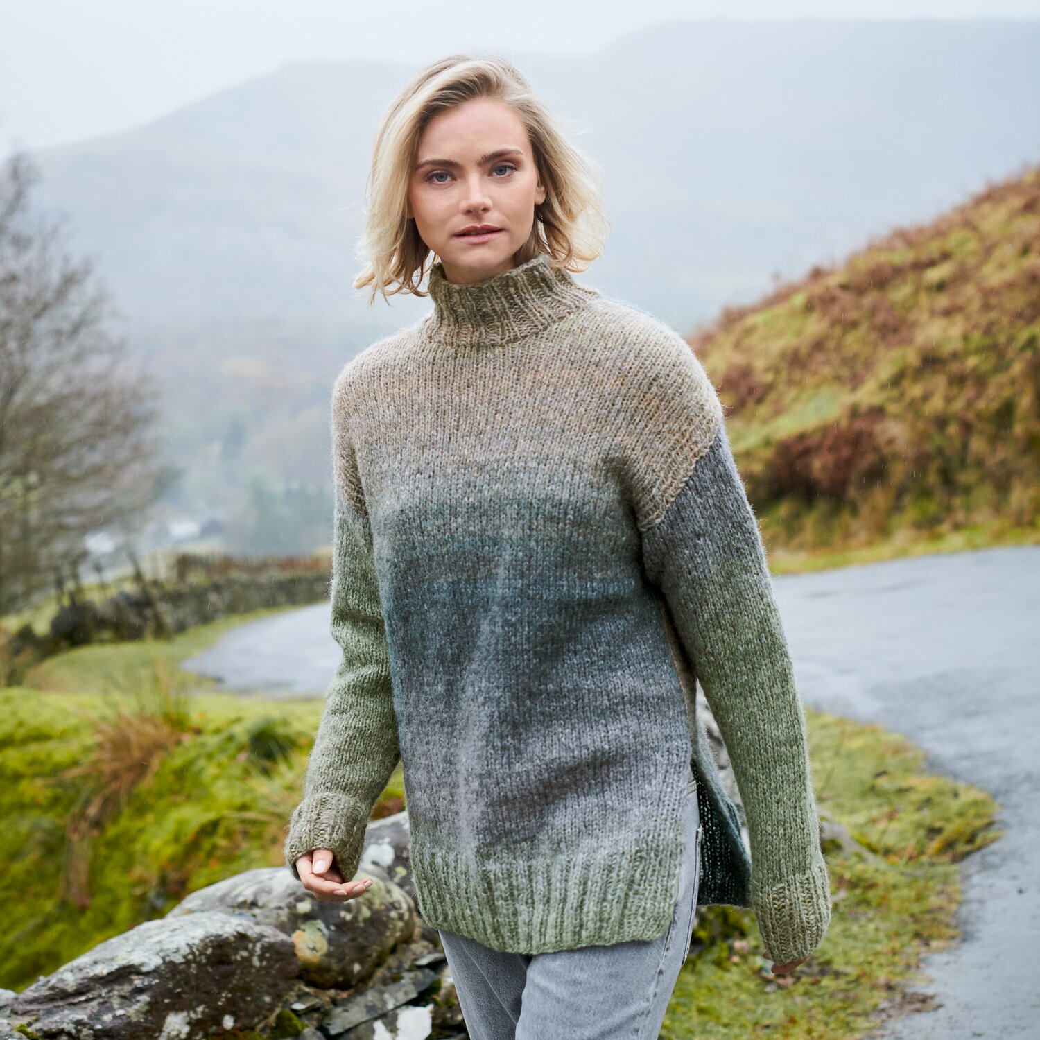 Strickset Pullover Modell 17 aus Lovewool No. 23, locker geschnitten mit hohem Kragen, im harmonischen Farbverlauf von Beige zu Grau, getragen von Frau in Landschaft