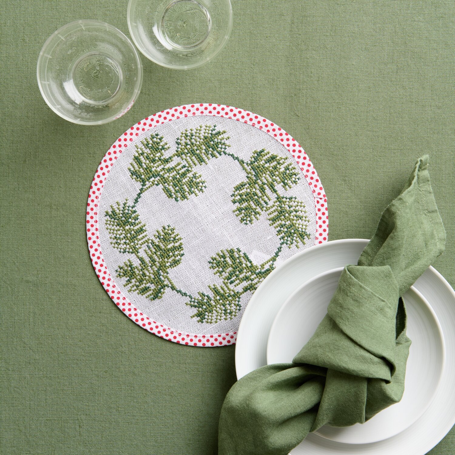 Stickpackung Untersetzer rund Tannenzweige zeigt einen runden Untersetzer mit Tannenzweig-Muster auf grünem Tischtuch, umgeben von Geschirr.