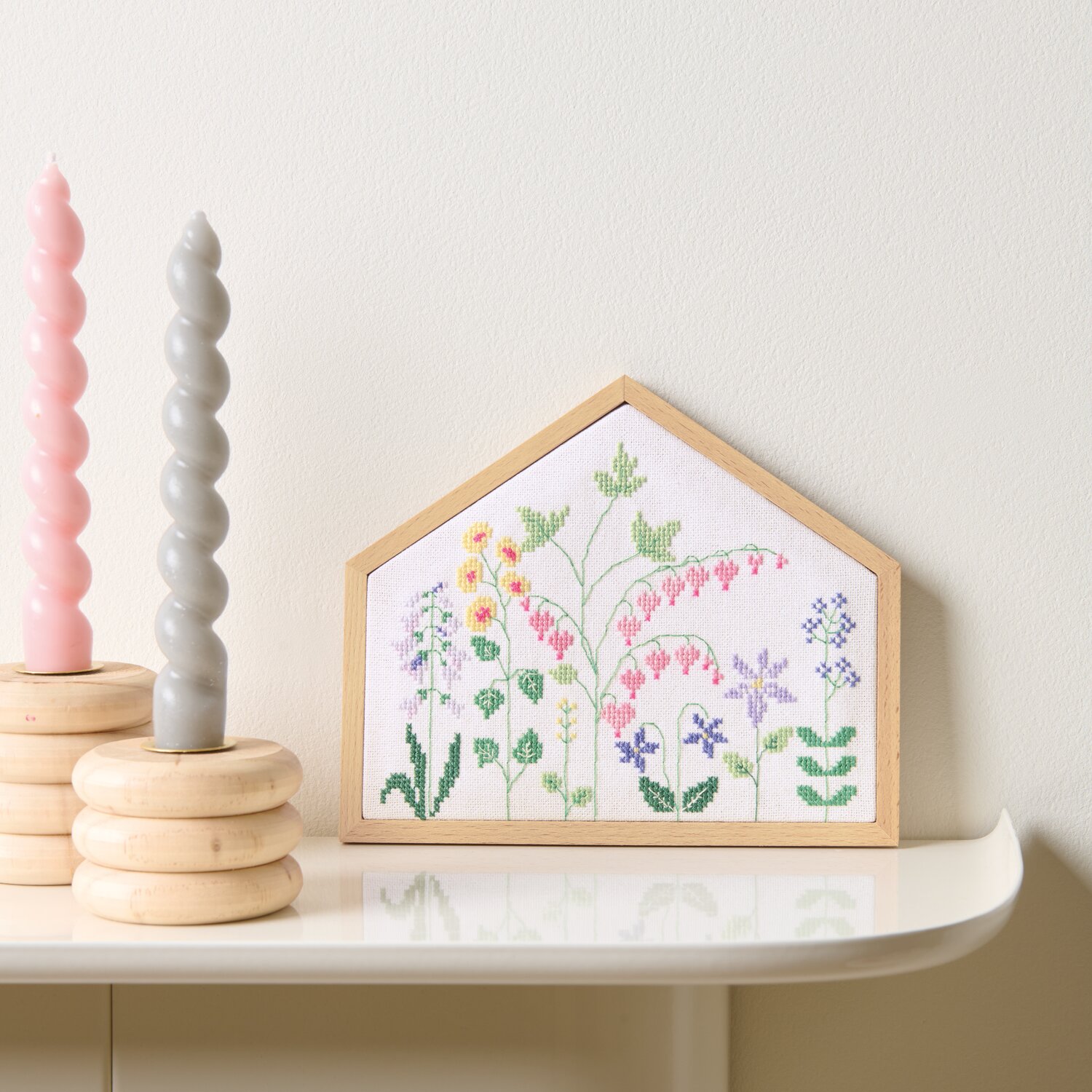 Stickpackung Gezählter Kreuzstich Blumenwiese zeigt bunte Blumen in Holzrahmen, dekoriert mit pastellfarbenen Kerzen auf weißem Regal.