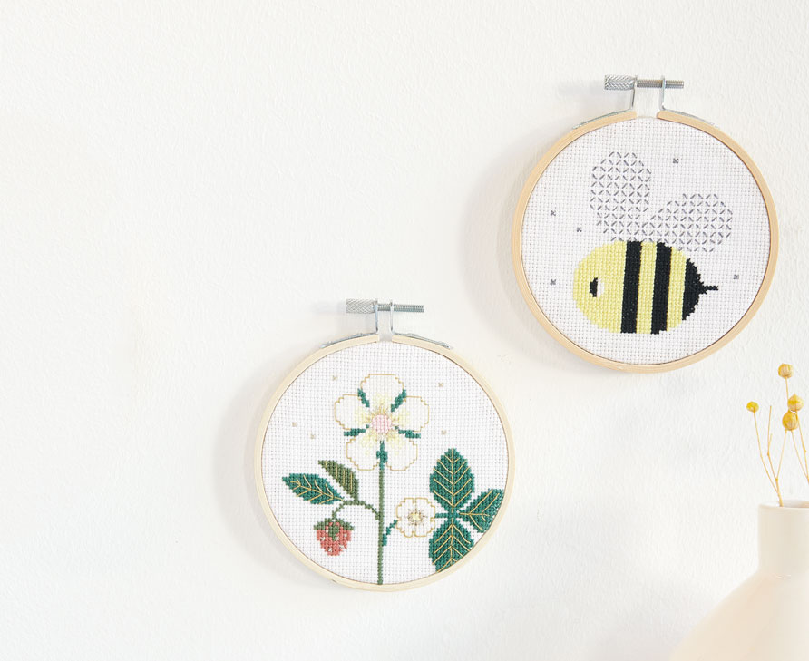 3 frühlingshafte Stickbilder im runden Stickrahmen mit den Motiven Erbeere und Biene sind an einer weißen Wand dekorativ angeordnet.