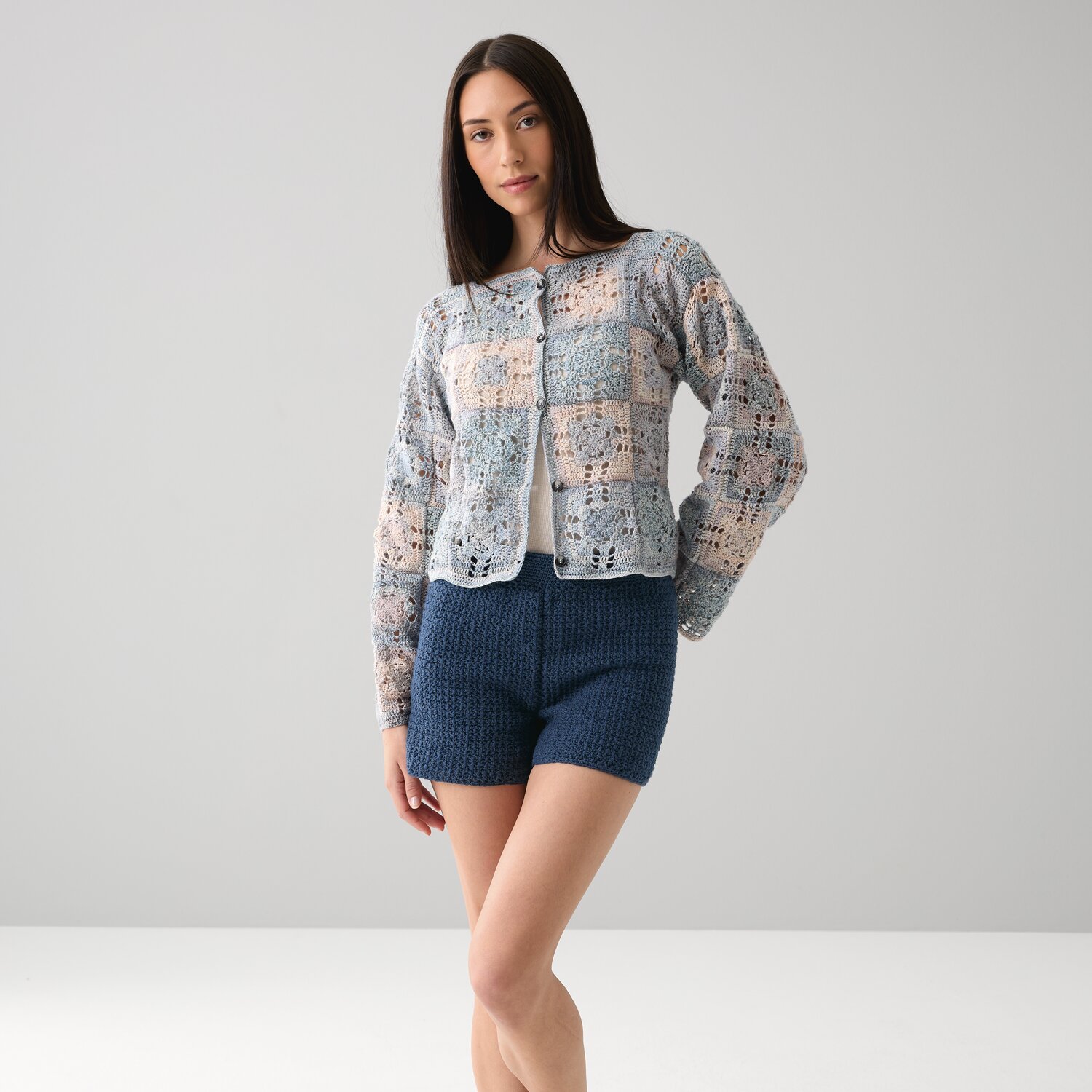 Häkelset Jacke Modell 07 aus Die Neue Masche No. 15, getragen mit blauen Häkelshorts, vor grauem Hintergrund.