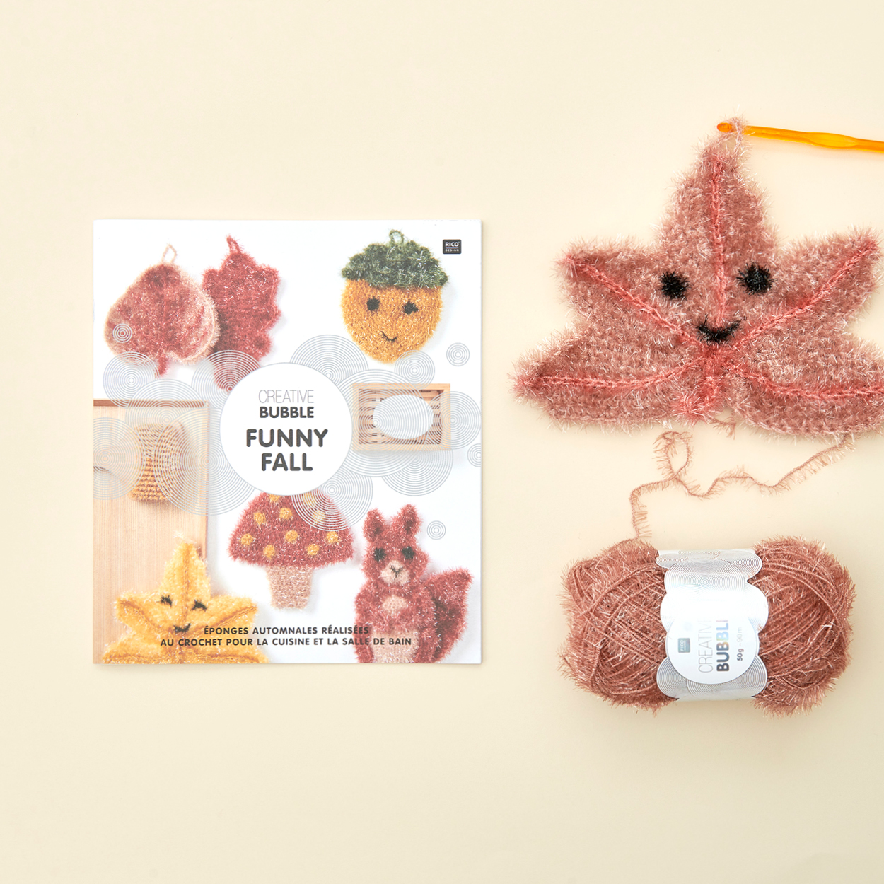 Creative Bubble Funny Fall Anleitungsheft daneben ein gehäkelter Schwamm in Blatt-Form mit Garn und Häkelnadel. Flatlay von Produkten.