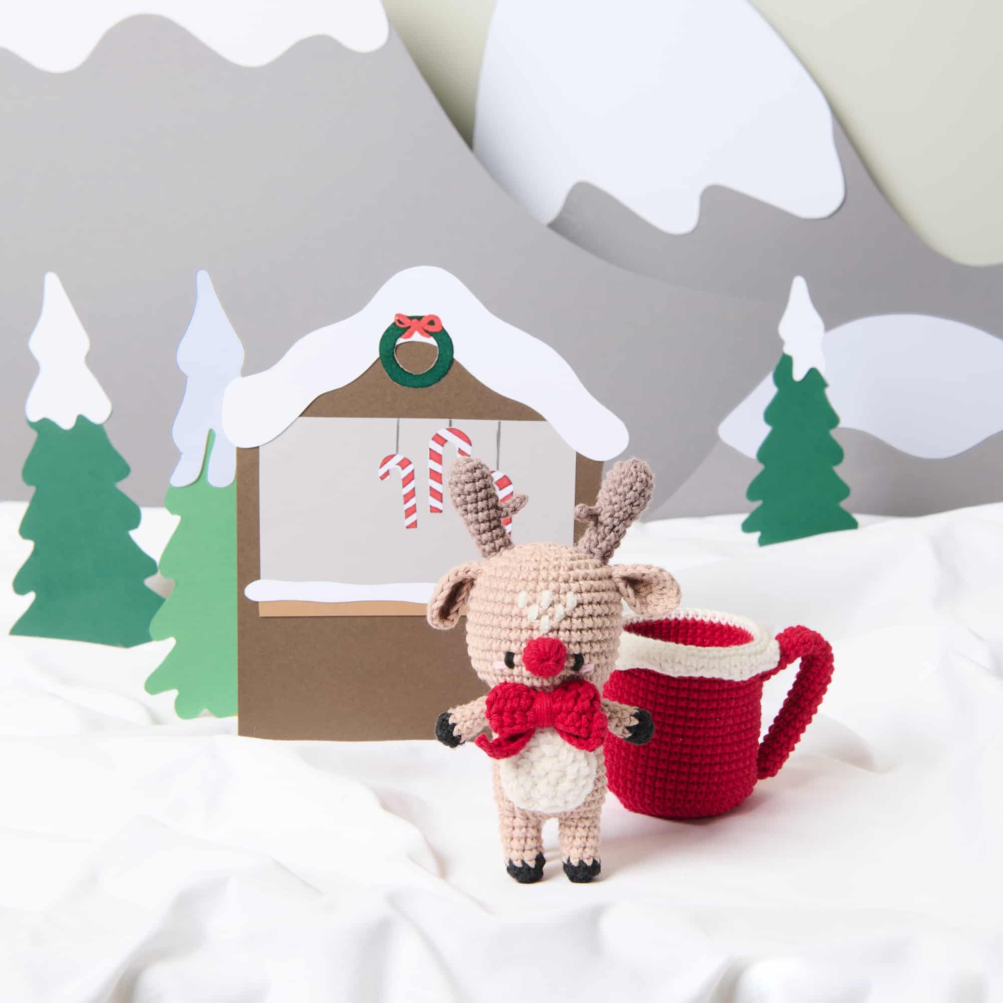 Ein gehäkeltes Rentier mit roter Tasse steht in einer Winter-Landschaft.