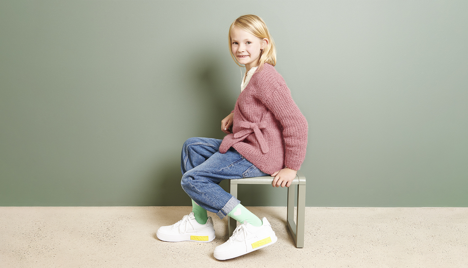 Strickanleitung Wickeljacke aus Soft Alpaca Strickanleitung Wickeljacke aus Soft Alpaca: Kind in rosa Jacke, Jeans und weißen Sneakers sitzt auf einer Bank vor grünem Hintergrund.