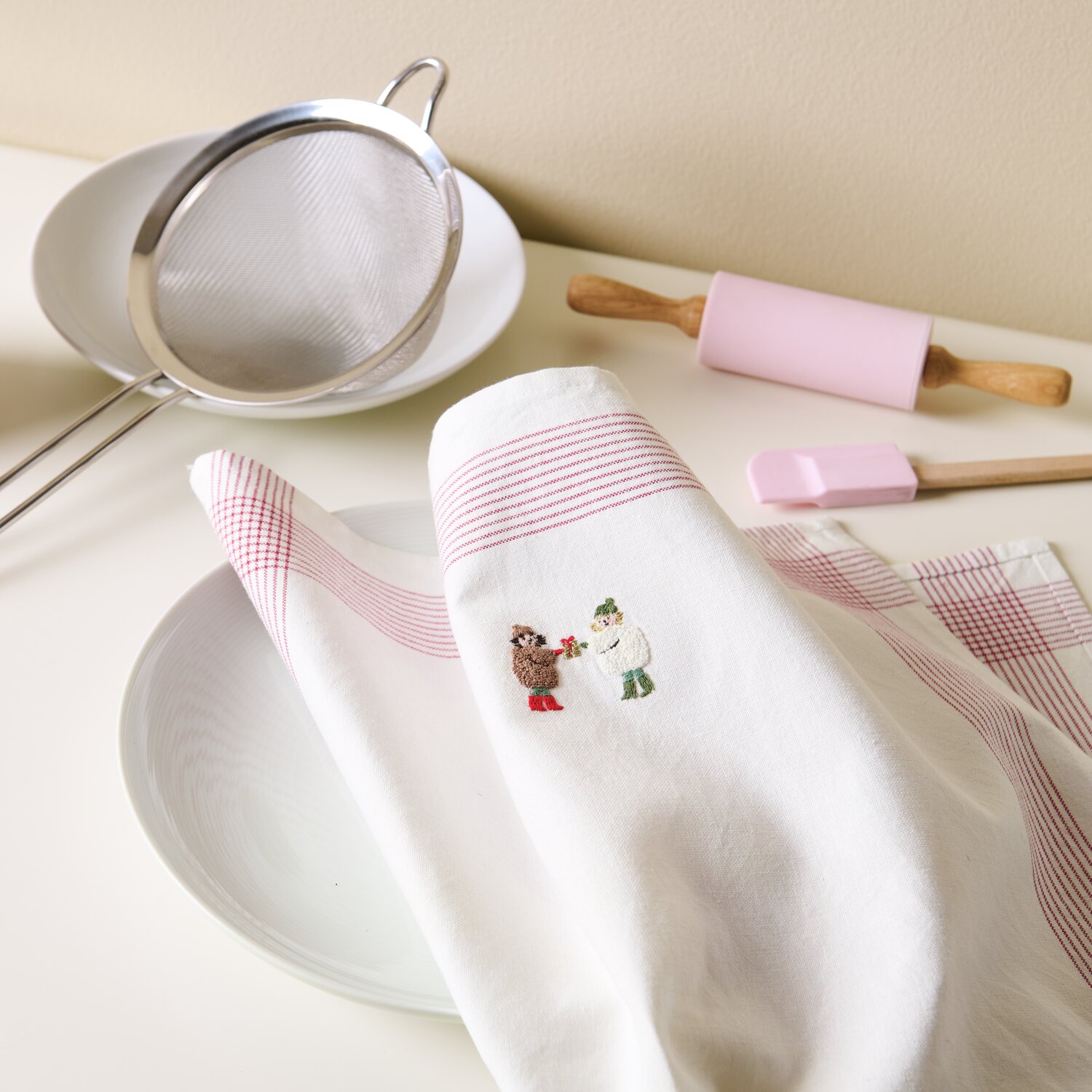 Stick & Stitch Stickpackung Kinder 15 Stück zeigt ein weißes Küchentuch mit rotem Muster und Stickerei. Im Hintergrund sind Küchenutensilien zu sehen.