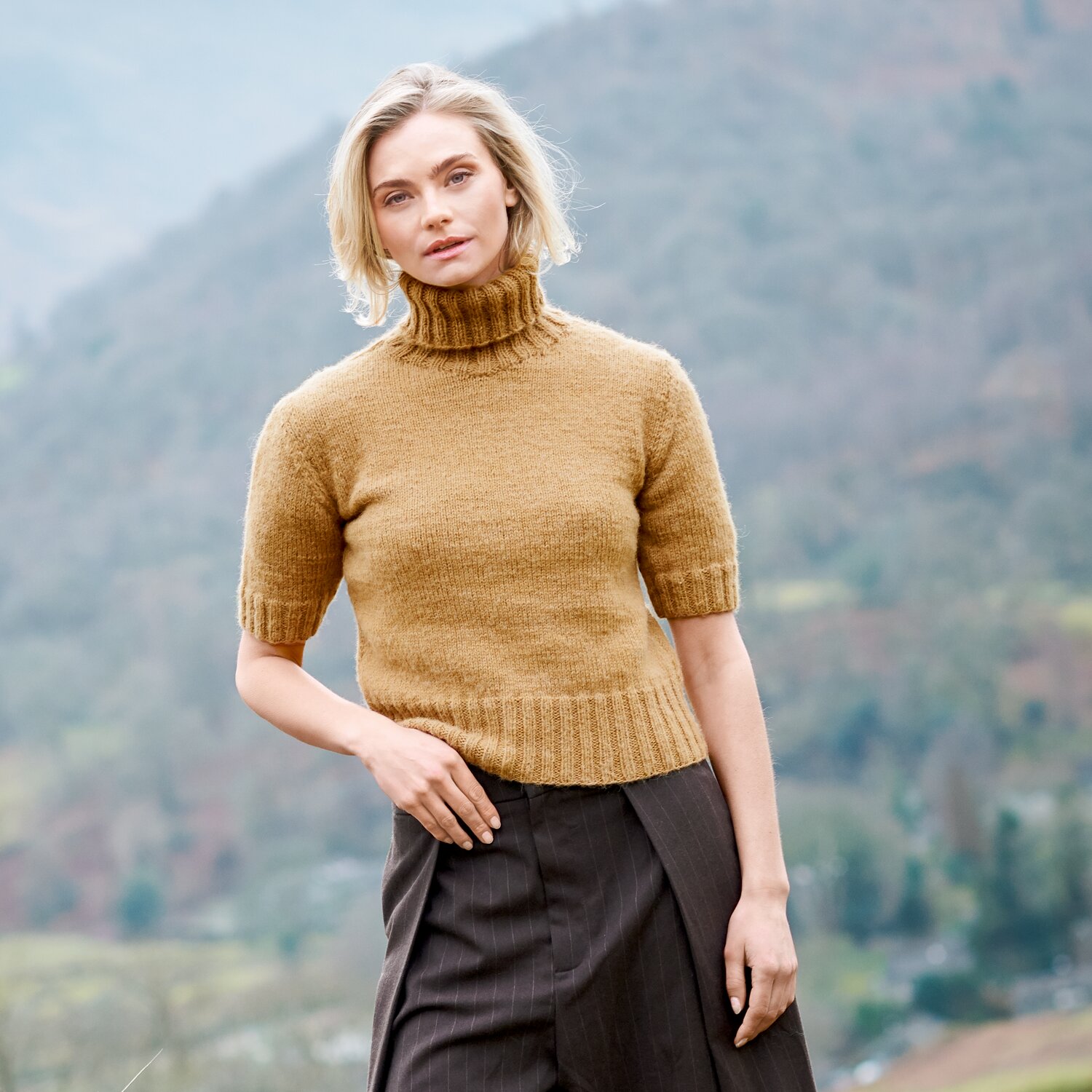 Strickset Shirt Modell 01 aus Lovewool No. 23, kurzärmeliges Rollkragenpullover-Modell in warmem Camelton, getragen von Frau vor bergiger Landschaft