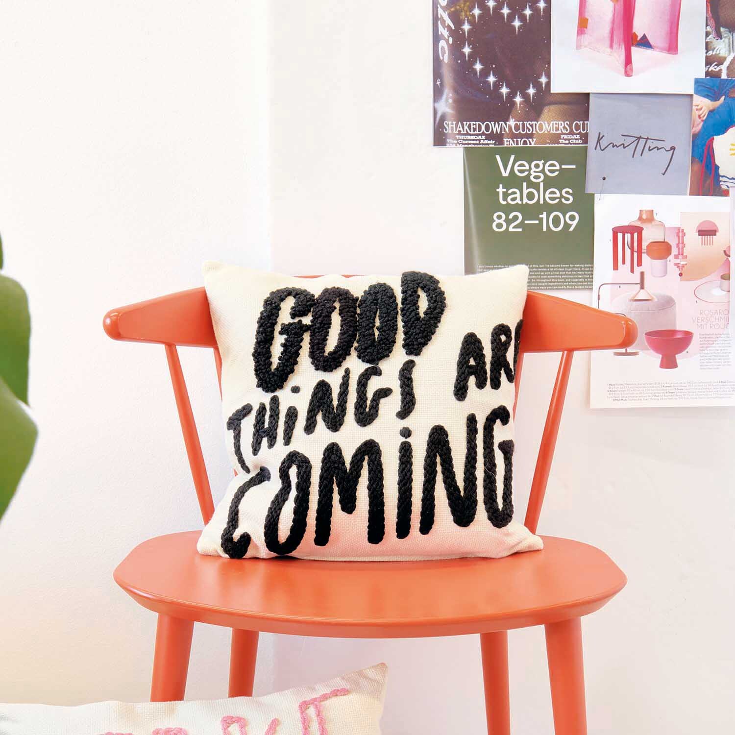 Anleitung Punch Needle Kissen mit Spruch auf einem orangefarbenen Stuhl, umgeben von Postern.