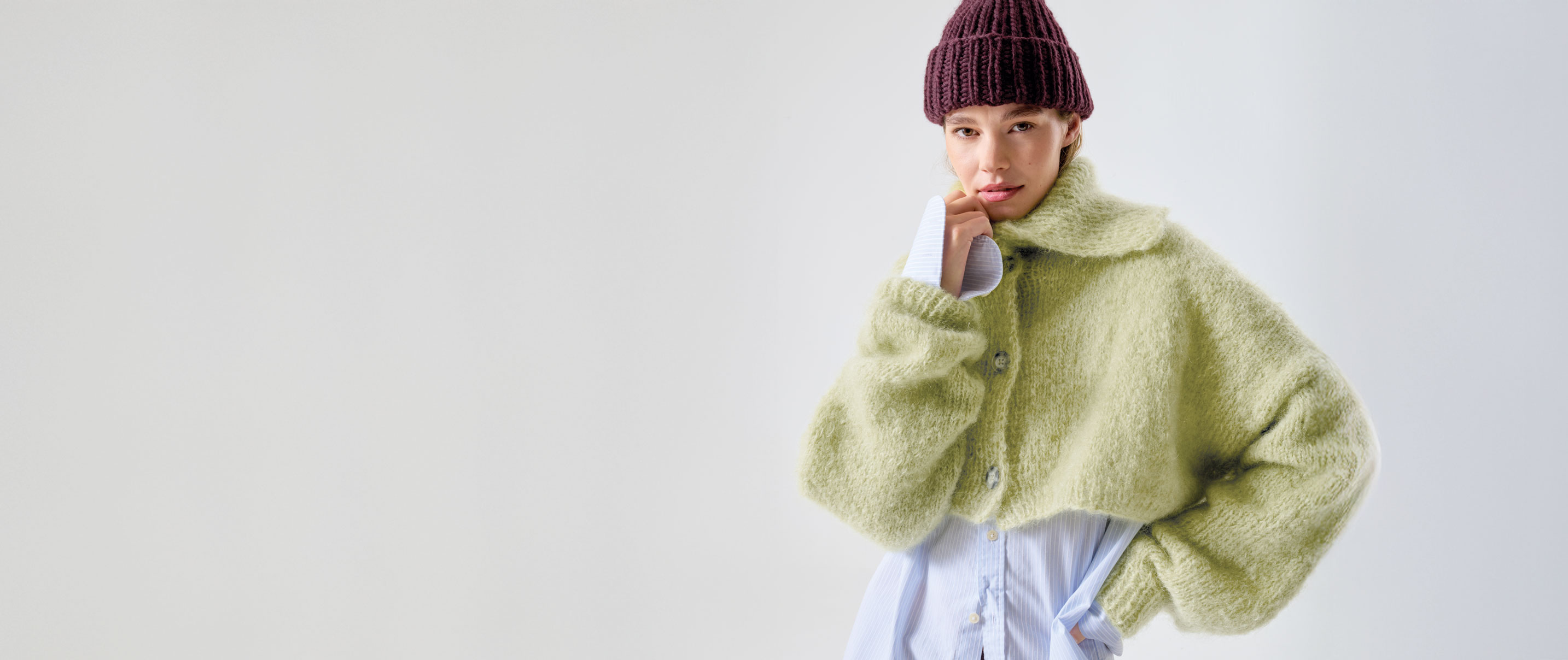 Frau trägt Strickjacke in grün und Mütze in aubergine vor hellem Hintergrund