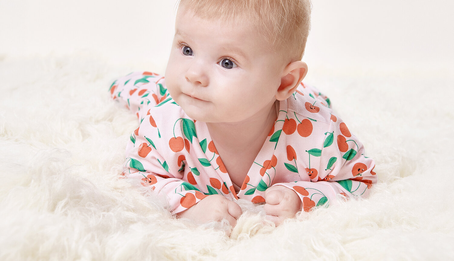 Nähanleitung Babystrampler aus Jerseystoff, liegendes Baby im Overall mit Kirschmuster auf weicher, heller Flauschdecke, Fokus auf gemütlicher Passform