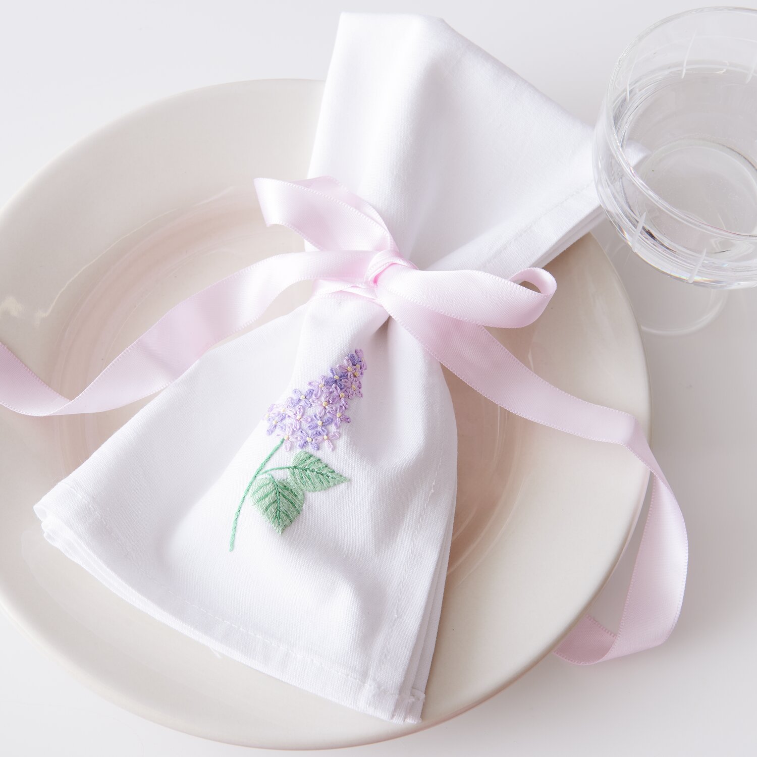 Stick & Stitch Packung Blumen XL 8 Stück: Weißes Tuch mit lila Blumenstickerei, rosa Schleife, auf Teller.