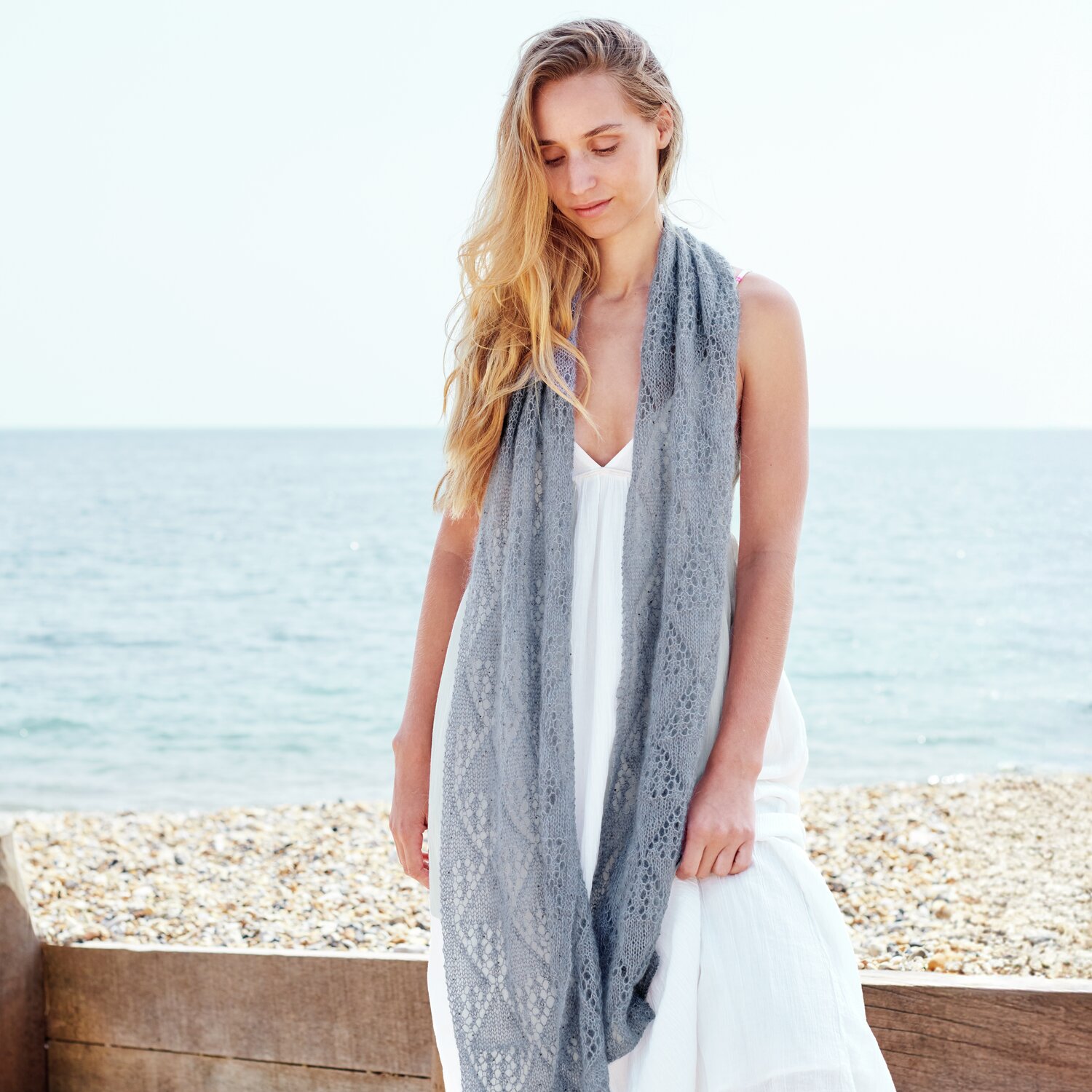 Strickset Tuch Modell 30 aus Lovewool No. 22: Frau am Strand mit langem, grauem Tuch über weißem Kleid.