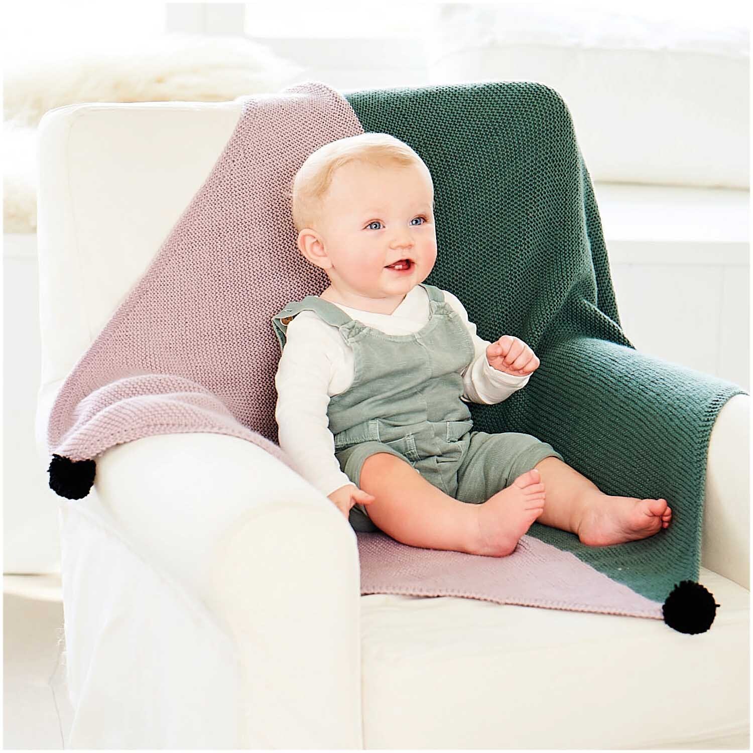 Rico Baby Dream dk uni - A Luxury Touch: Ein Baby sitzt auf einem Sessel mit gestrickter Decke in Rosa und Grün.
