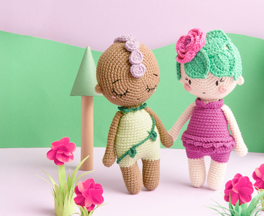 Ricorumi Häkelfiguren in Form von Blumen stehen vor gebasteltem Hintergrund und halten Händchen.