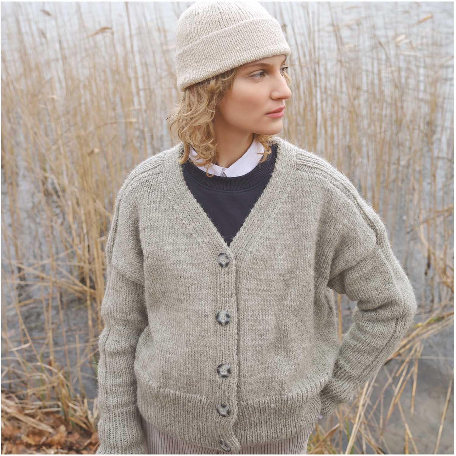 Strickset Mütze Modell 09 aus Luxury Knits Winter Special Marine: Frau in grauem Strickcardigan und beiger Mütze am Seeufer.