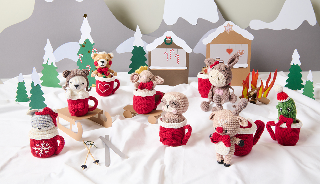 Häkelanleitung Ricorumi CAL Daisy the Christmas Cookie Donkey: Weihnachtliche Amigurumi-Szene mit Tassen und Figuren in Rot und Beige vor winterlicher Kulisse.