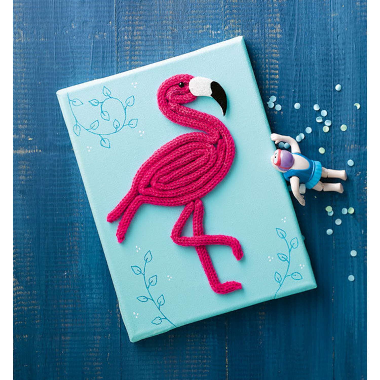Kreativ kinderleicht Strickliesel: Flamingo aus pinkem Garn auf hellblauer Leinwand, dekoriert mit blauen Blättern.