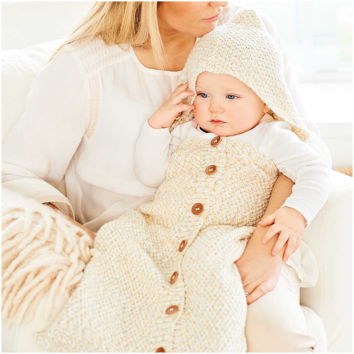 Rico Baby Dream dk A Luxury Touch: Baby in gestricktem Pastellkonfetti-Schlafsack auf Sofa mit Mutter.