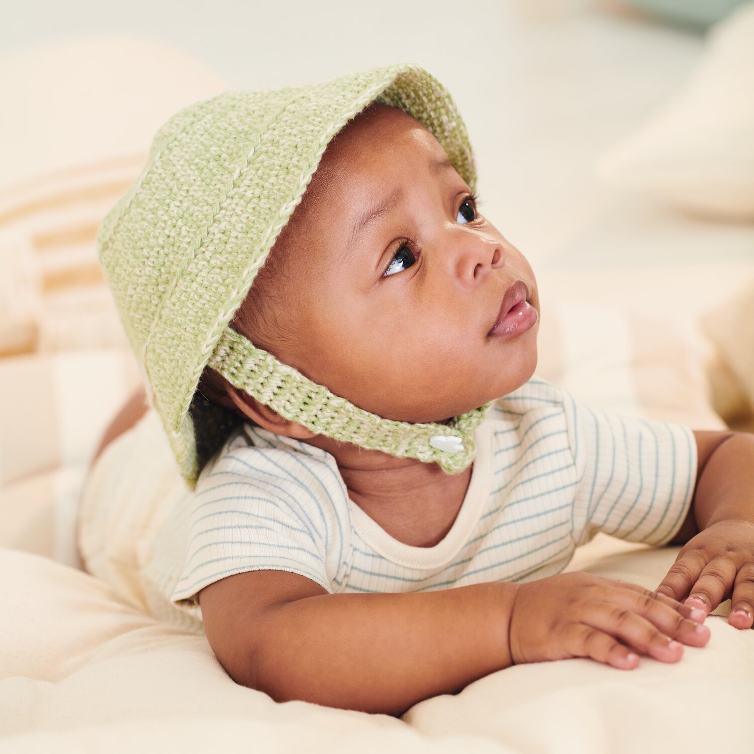 Häkelset Mütze Modell 12b aus Baby 043: Baby mit grüner Häkelmütze, liegend auf beigem Untergrund.