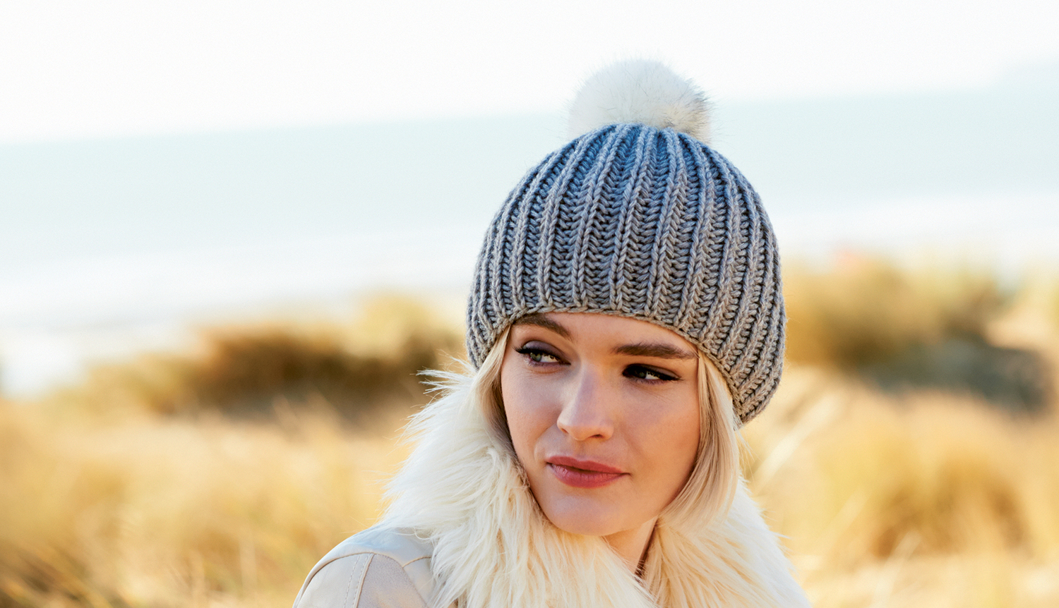 Strickanleitung Mütze mit Kunstfellpompon Strickanleitung Mütze mit Kunstfellpompon: Frau trägt graue Strickmütze mit weißem Pompon am Strand.