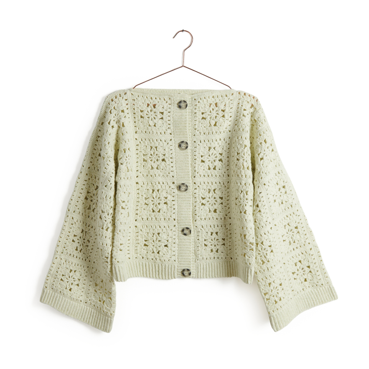 Eine Jacke mit Knöpfen aus einem Cremefarbenden Garn, gehäkelt hängt an einem Kleiderbügel.