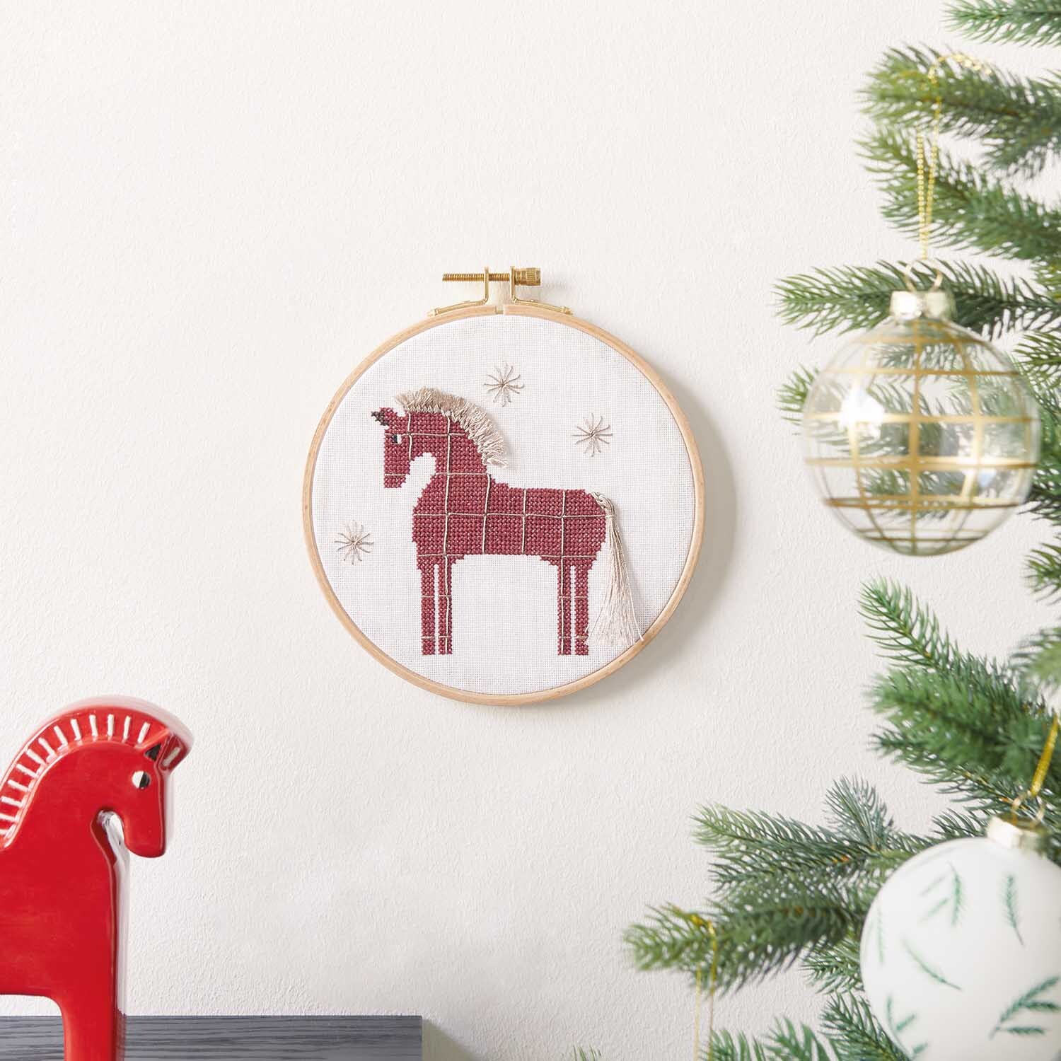 Stickanleitung Pferdemotiv im Stickring: Rotes Pferd im Stickrahmen an weißer Wand, dekoriert mit Weihnachtsbaum.