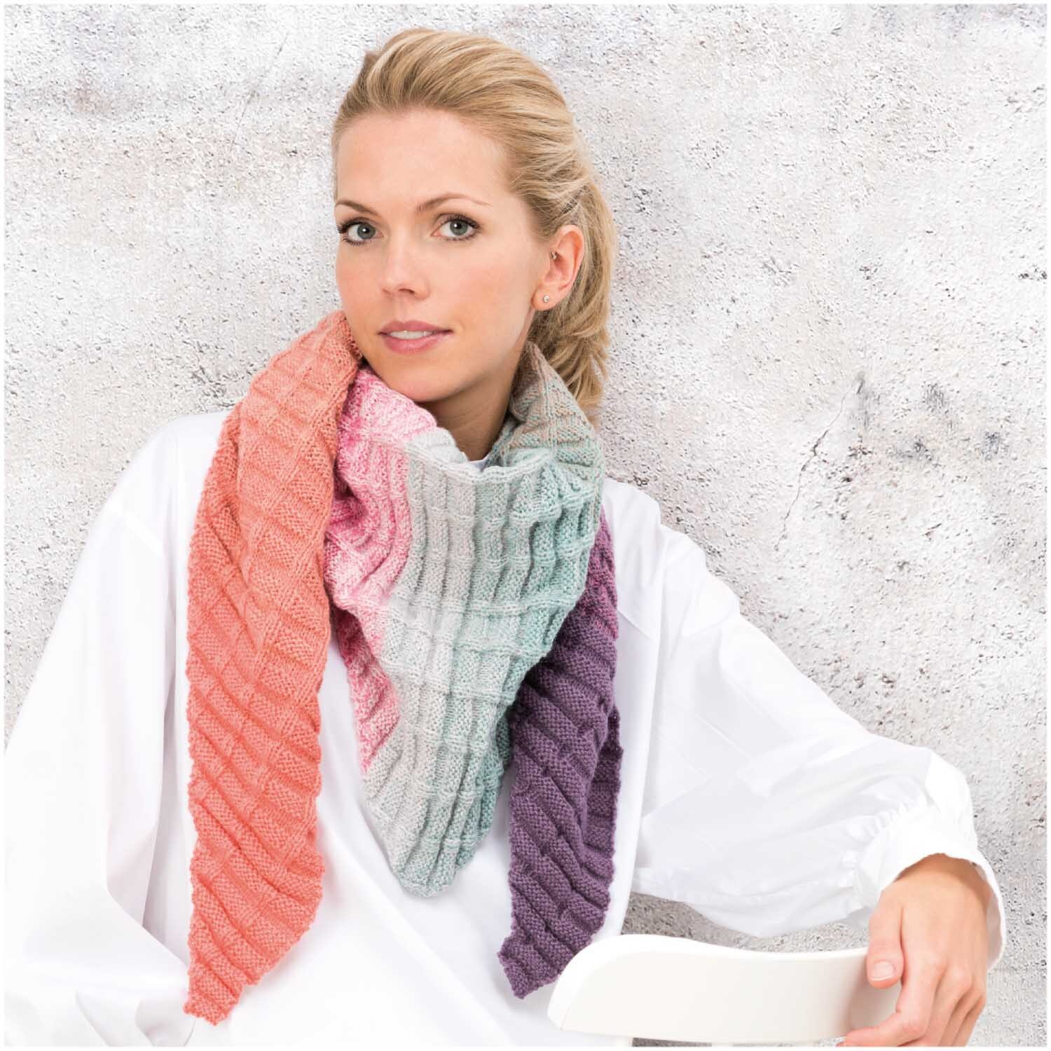 Creative Wool Dégradé Super6: Frau trägt gestrickten Schal in Regenbogenfarben von Rico Design vor grauer Wand.