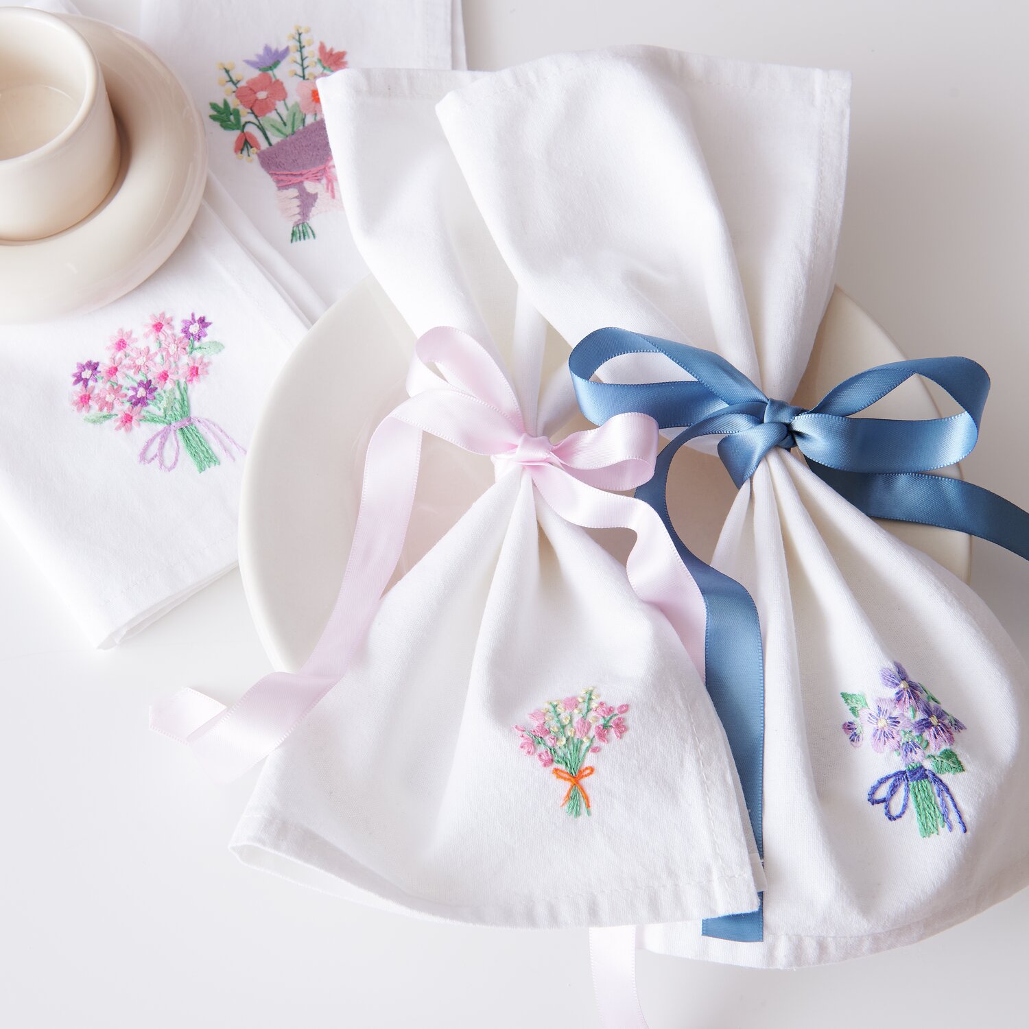 Stick & Stitch Packung Blumensträuße XL 7 Stück: Weiße Stoffservietten mit bunten Blumenstickereien und farbigen Schleifen.