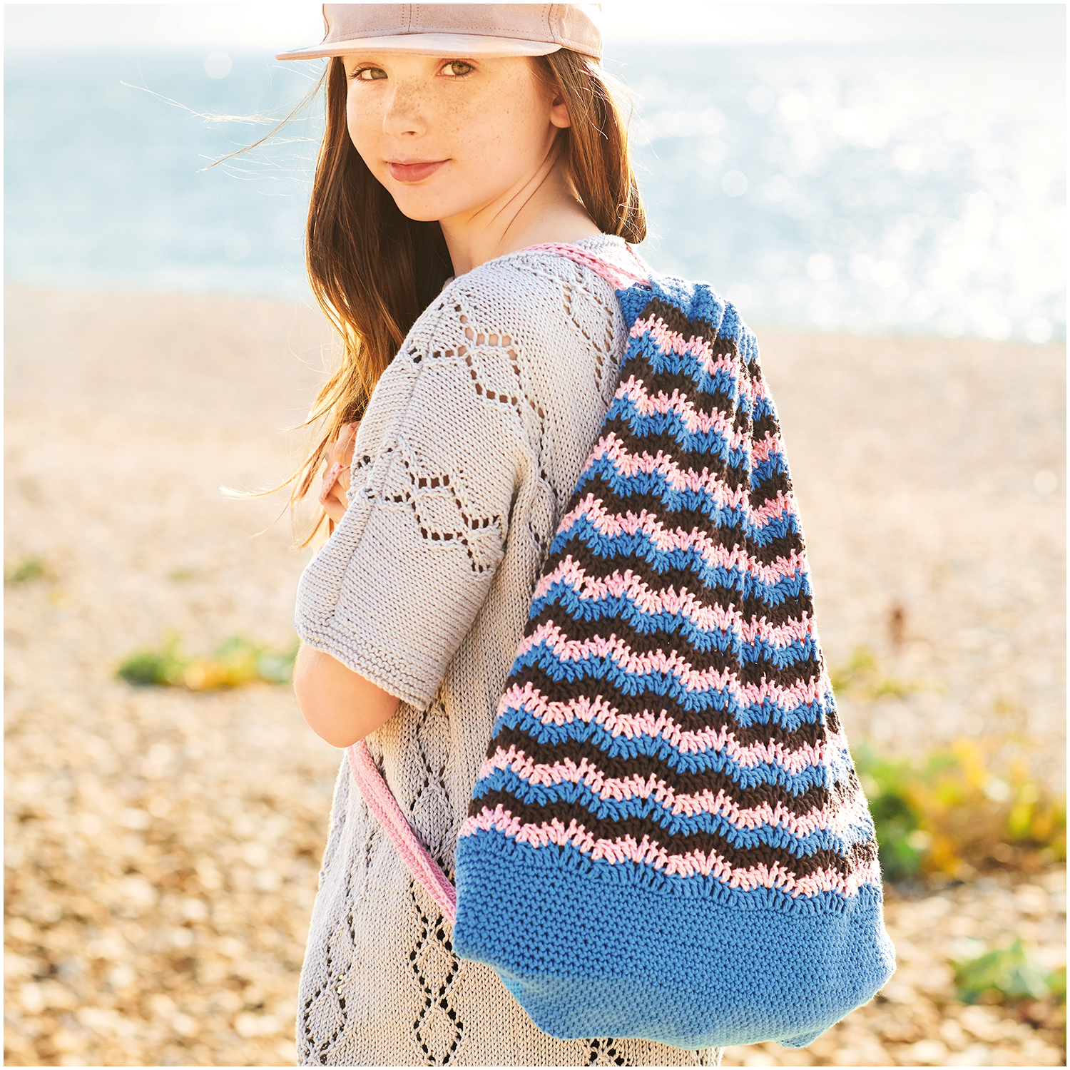 Essentials Organic Cotton aran: Mädchen am Strand mit gestricktem Rucksack in Blau- und Rosatönen. Marke: Rico Design.