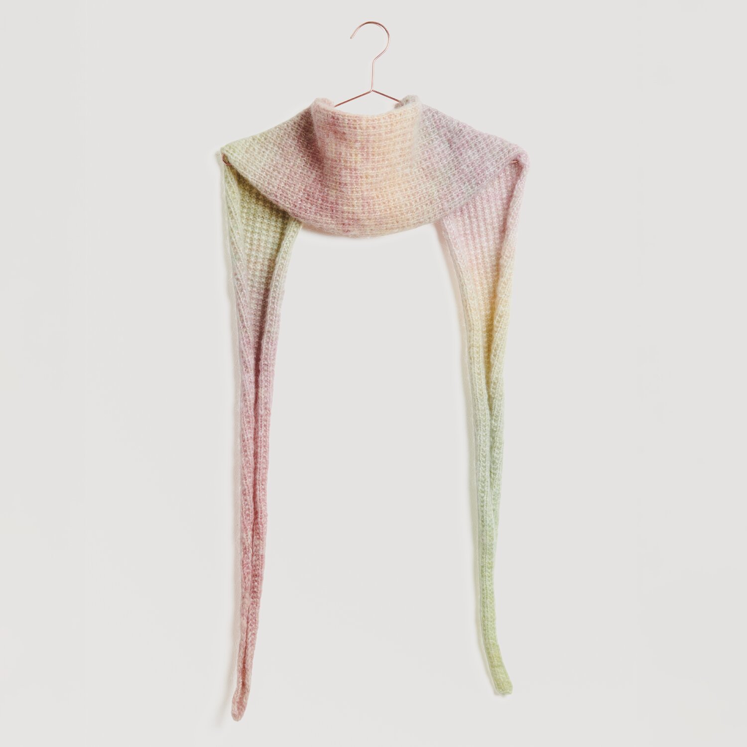 Ein langer, gestrickter Schal in sanften Pastelltönen hängt an einem Haken. Der Schal zeigt einen Farbverlauf von Rosa zu Grün. Marke: Modell Legeware. Ideal für stilvolle Wintermode.