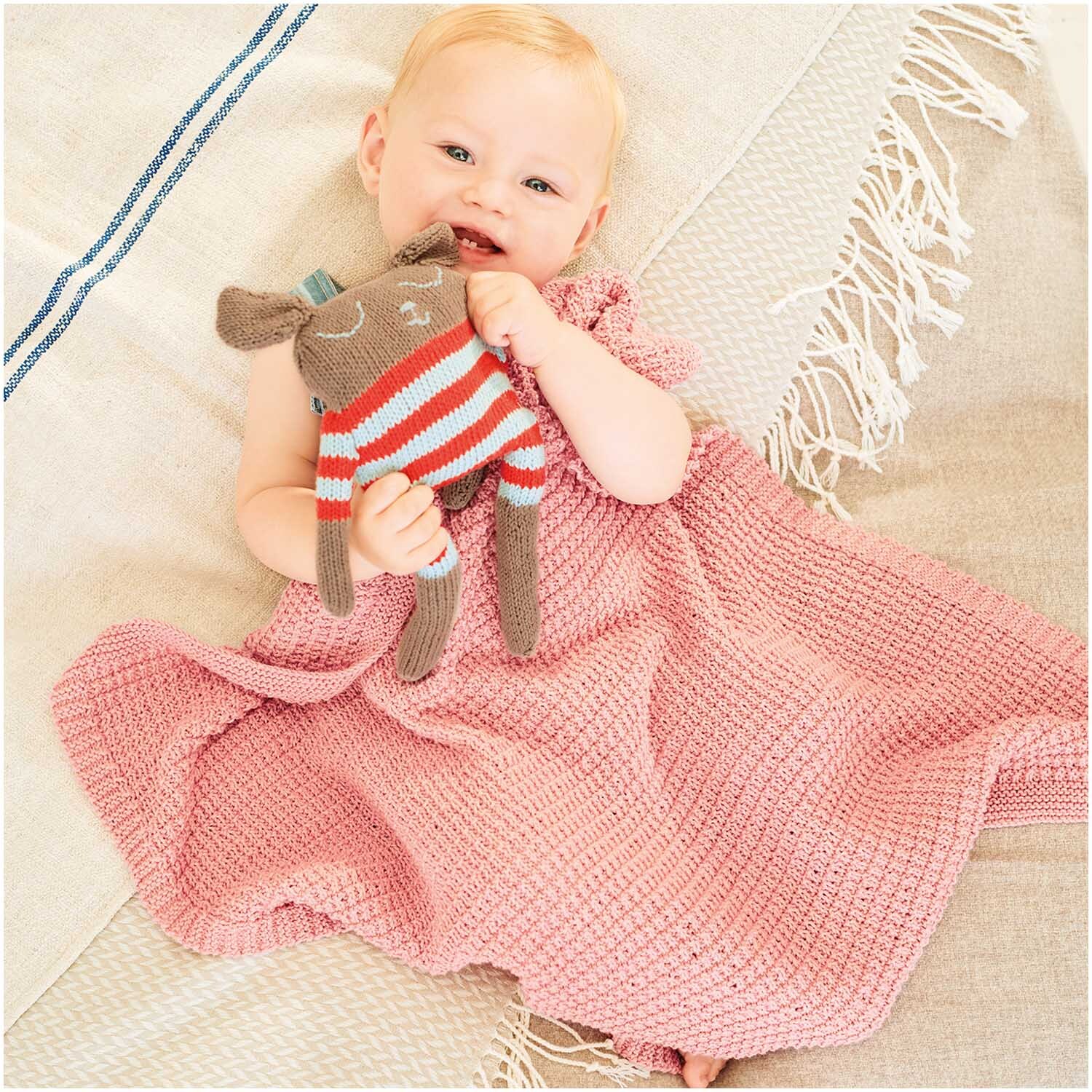 Baby Organic Cotton: Ein Baby in einem himbeerfarbenen Strickkleid hält ein gestricktes Stofftier.