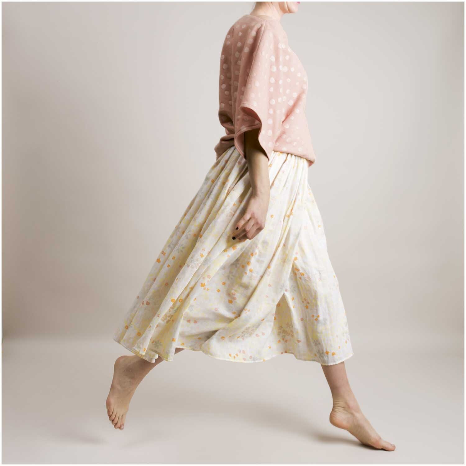 Musselin-Druckstoff Crafted Nature Blumenwiese Grau-Metallic 140cm: Frau in rosa Oberteil und geblümtem Rock schreitet barfuß vor neutralem Hintergrund.