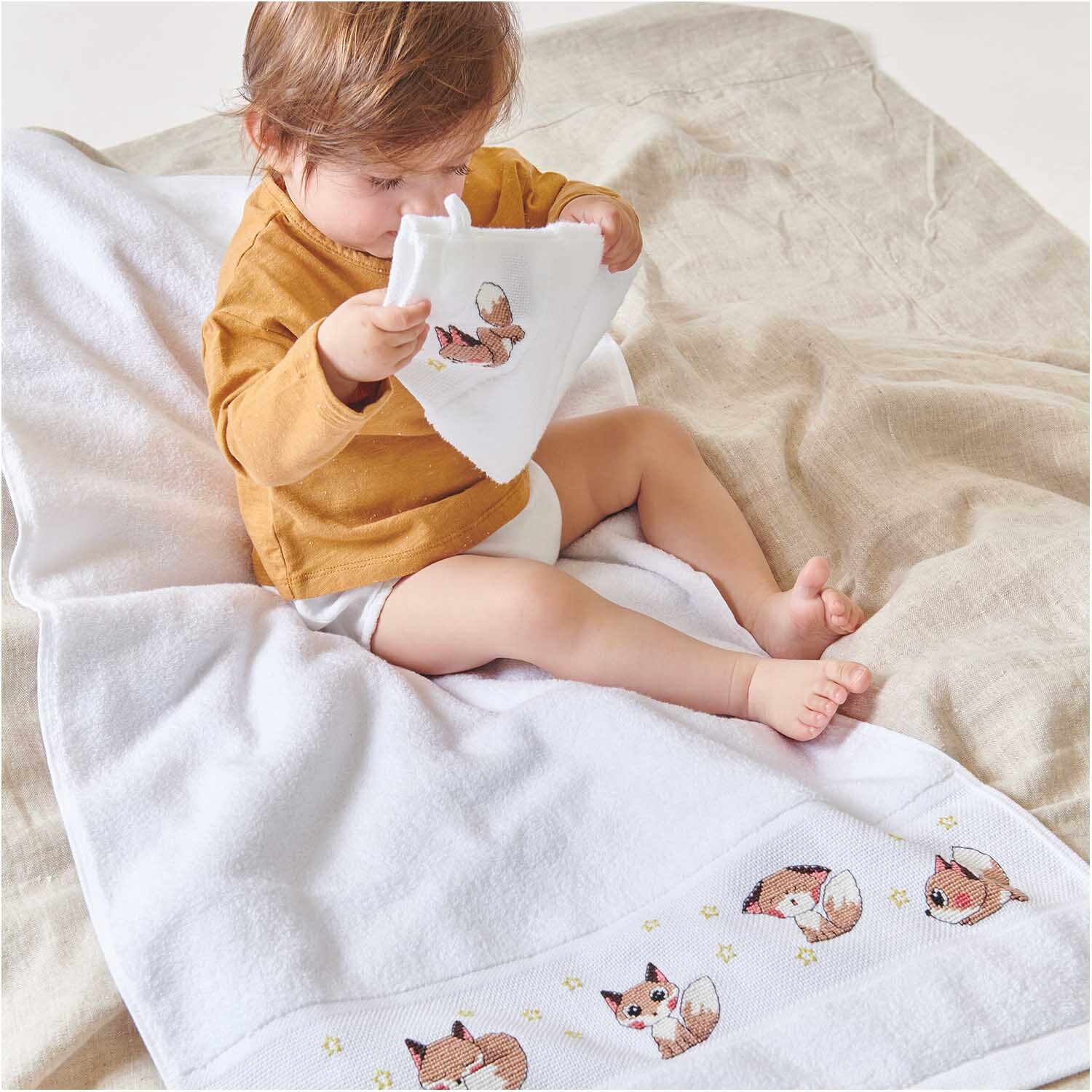 Handtuch 50x100cm Farbe Weiß von Rico Design Handtuch 50x100cm: Ein Kind sitzt auf einem weißen Handtuch mit Fuchs-Motiven, trägt ein senfgelbes Oberteil. Ideal für Kinderzimmer.