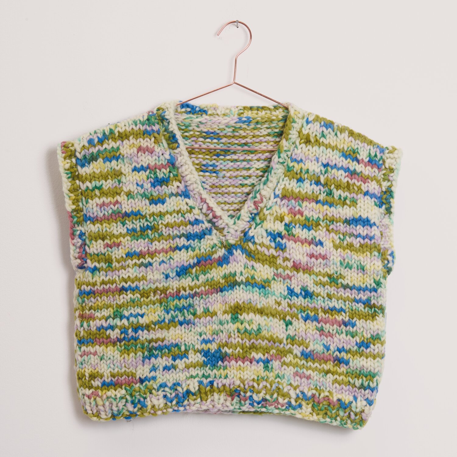 Fashion Tinted Merino Stricktop mit V-Ausschnitt aus mehrfarbigem Garn von Rico Design, locker gestrickt und auf Kupferbügel vor hellrosa Wand präsentiert