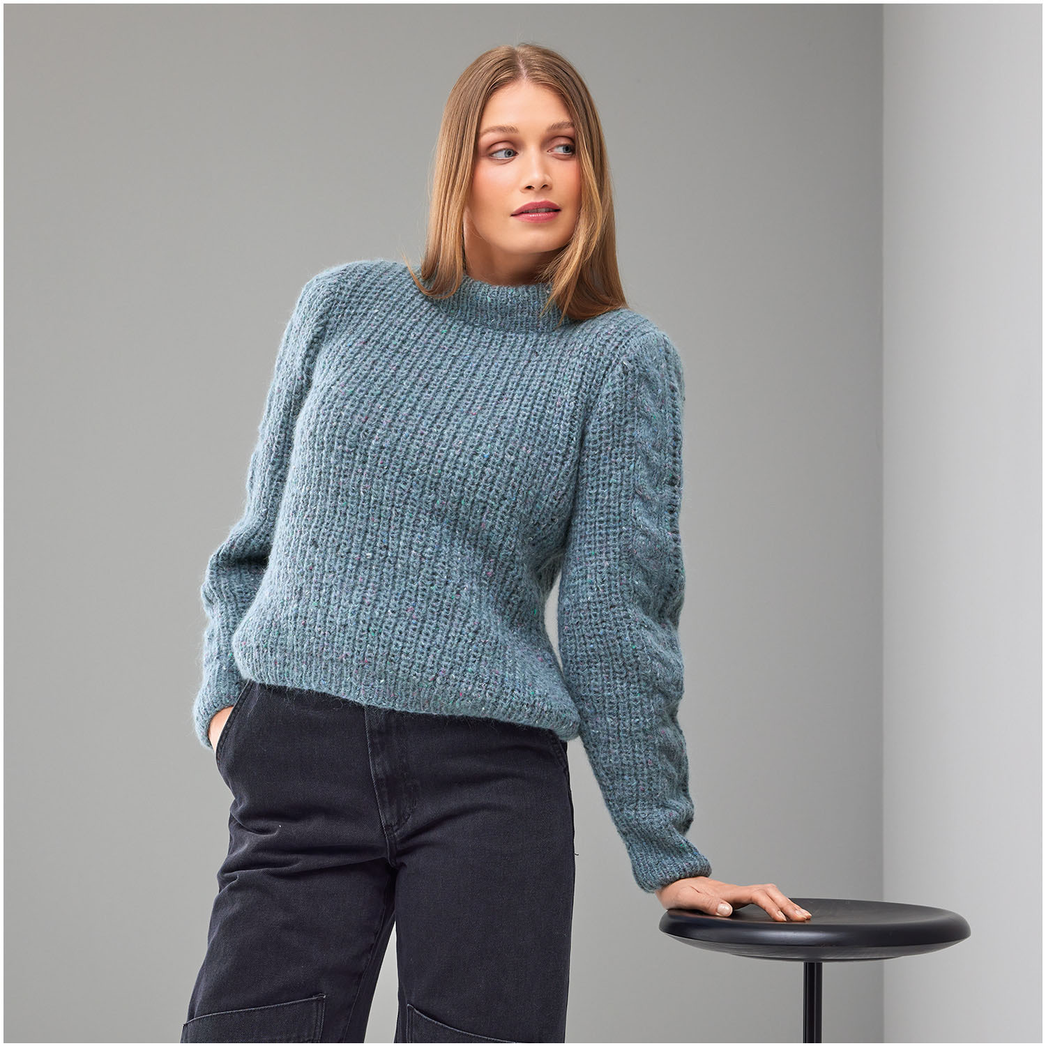 Strickset Pullover Modell 24 aus Die Neue Masche Nr. 8: Frau trägt blauen Pullover, lehnt an einem Tisch, grauer Hintergrund.