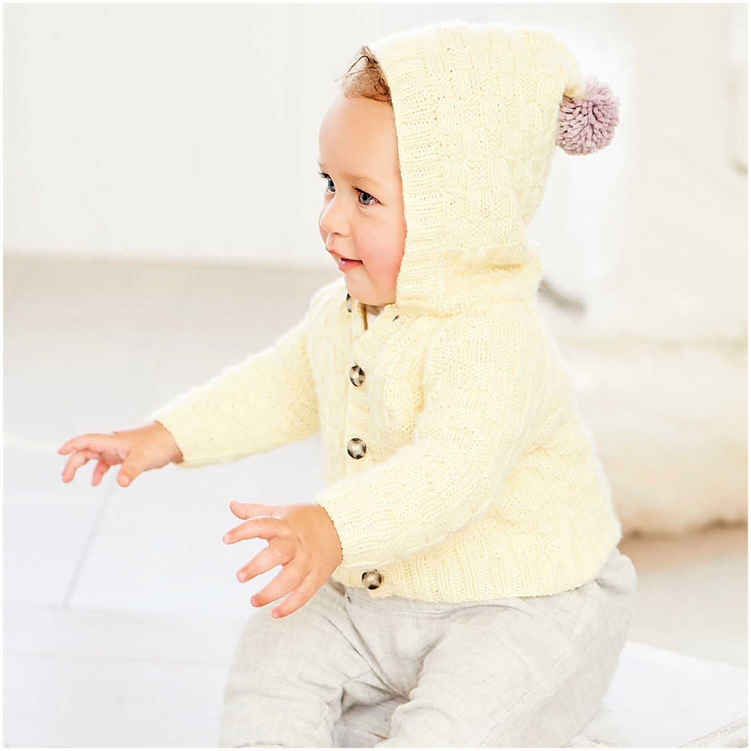 Rico Baby Dream dk uni - A Luxury Touch: Ein Baby trägt einen cremefarbenen Strickpullover mit Kapuze und Pompon.