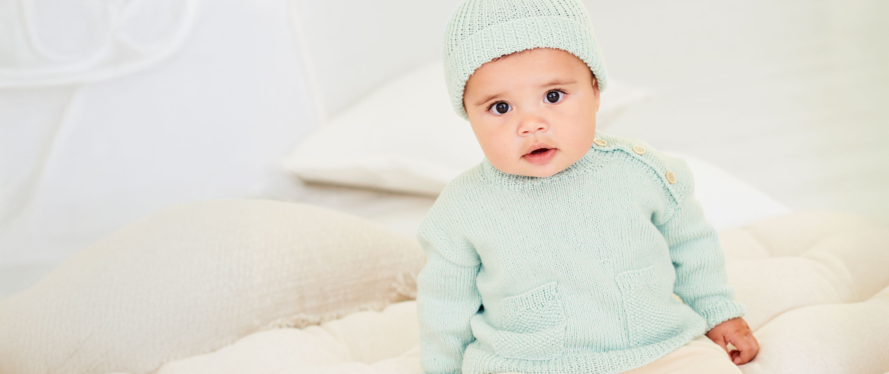 Baby trägt mintfarbenen Strickpullover und mintfarbene Strickmütze und sitzt auf Kissen und Decken.
