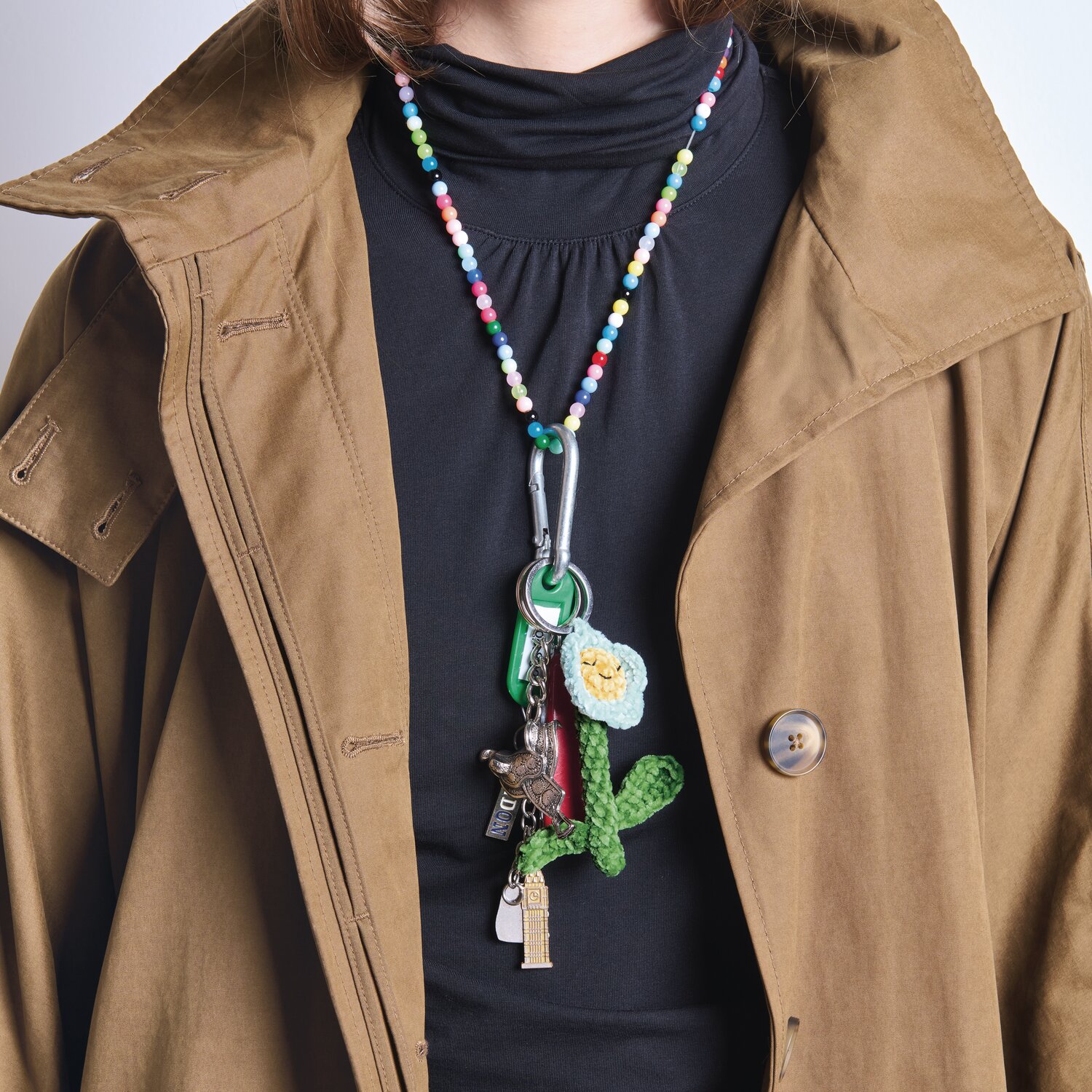 Ricorumi Häkelset Schlüsselanhänger Chingu Earth an bunter Perlenkette, getragen über braunem Mantel und schwarzem Oberteil.