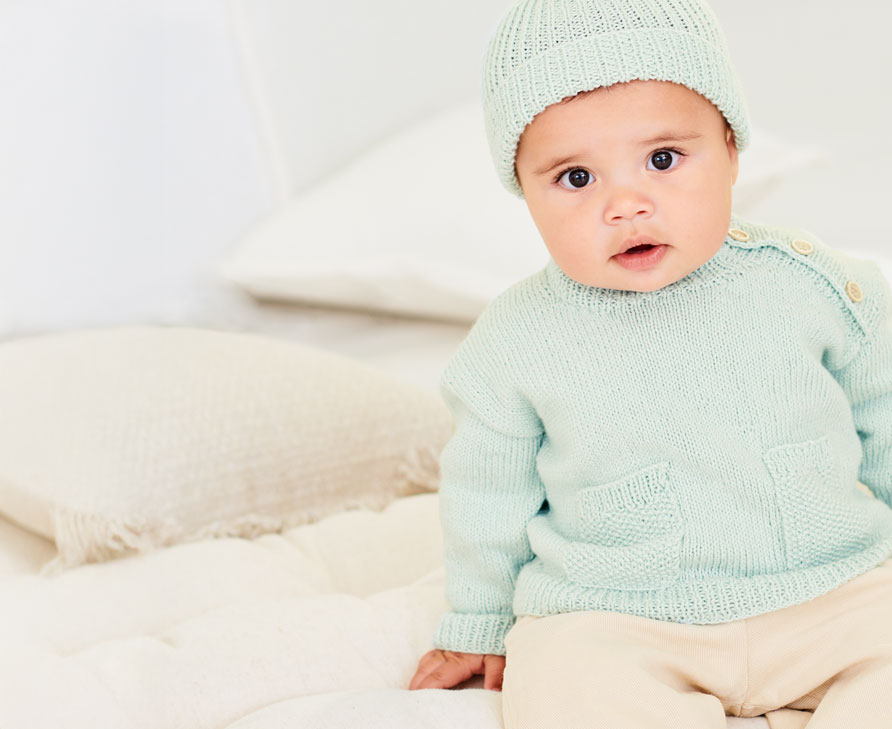 Baby trägt mintfarbenen Strickpullover und mintfarbene Strickmütze und sitzt auf Kissen und Decken.